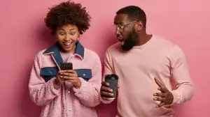 Emotive black woman and man stare at smartphone device, react on stunning news, get message, drink takeout coffee, have surprised expressions, dressed in casual wear. People and lifestyle concept