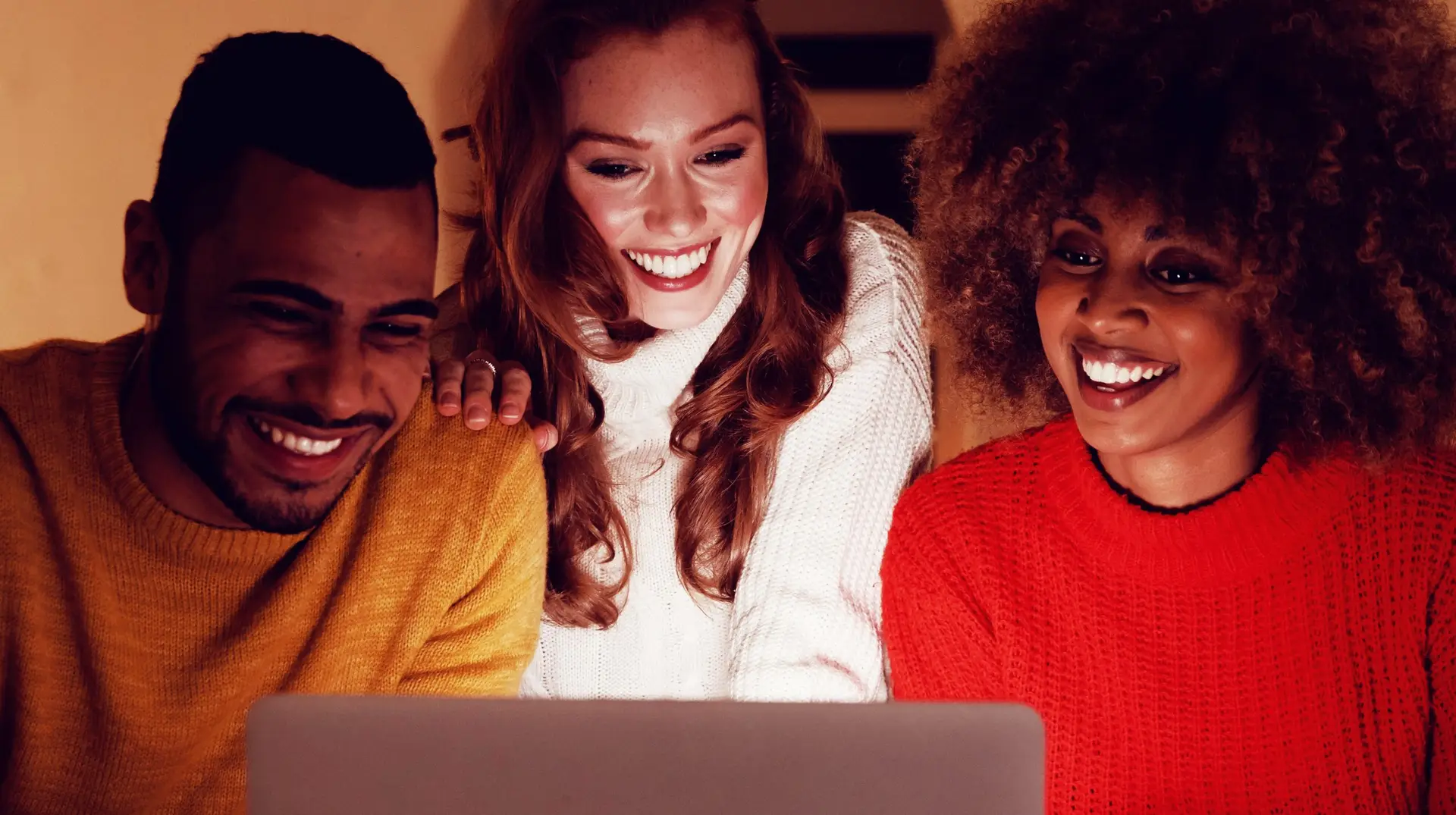 Front view of a young mixed race man and woman sitting at a table with a young Caucasian woman standing behind them, looking at a laptop computer together and smiling in the sitting room of an apartment