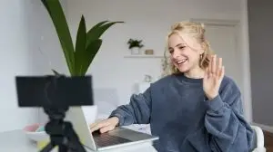 Portrait of young social media content creator, woman sits in room with laptop and digital camera, records lifestyle vlog, says hello, chats to someone online via computer