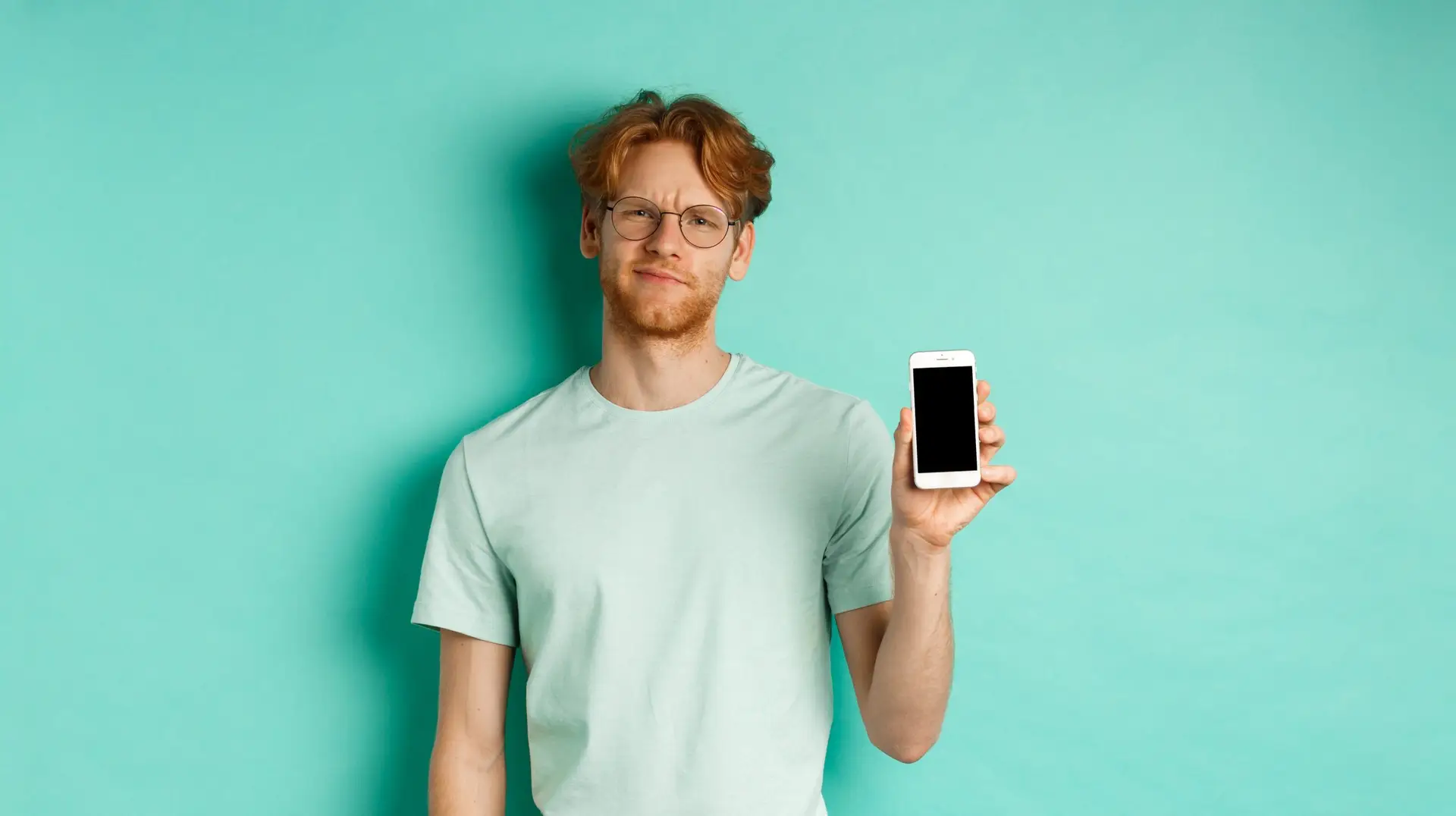 Skeptical male model with red hair and glasses, showing mobile screen and frowning disappointed, dislike application, standing over turquoise background