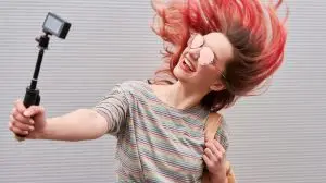 Young blogger woman with pink dyed flying hairs dancing near the grey wall and making vlog by action camera