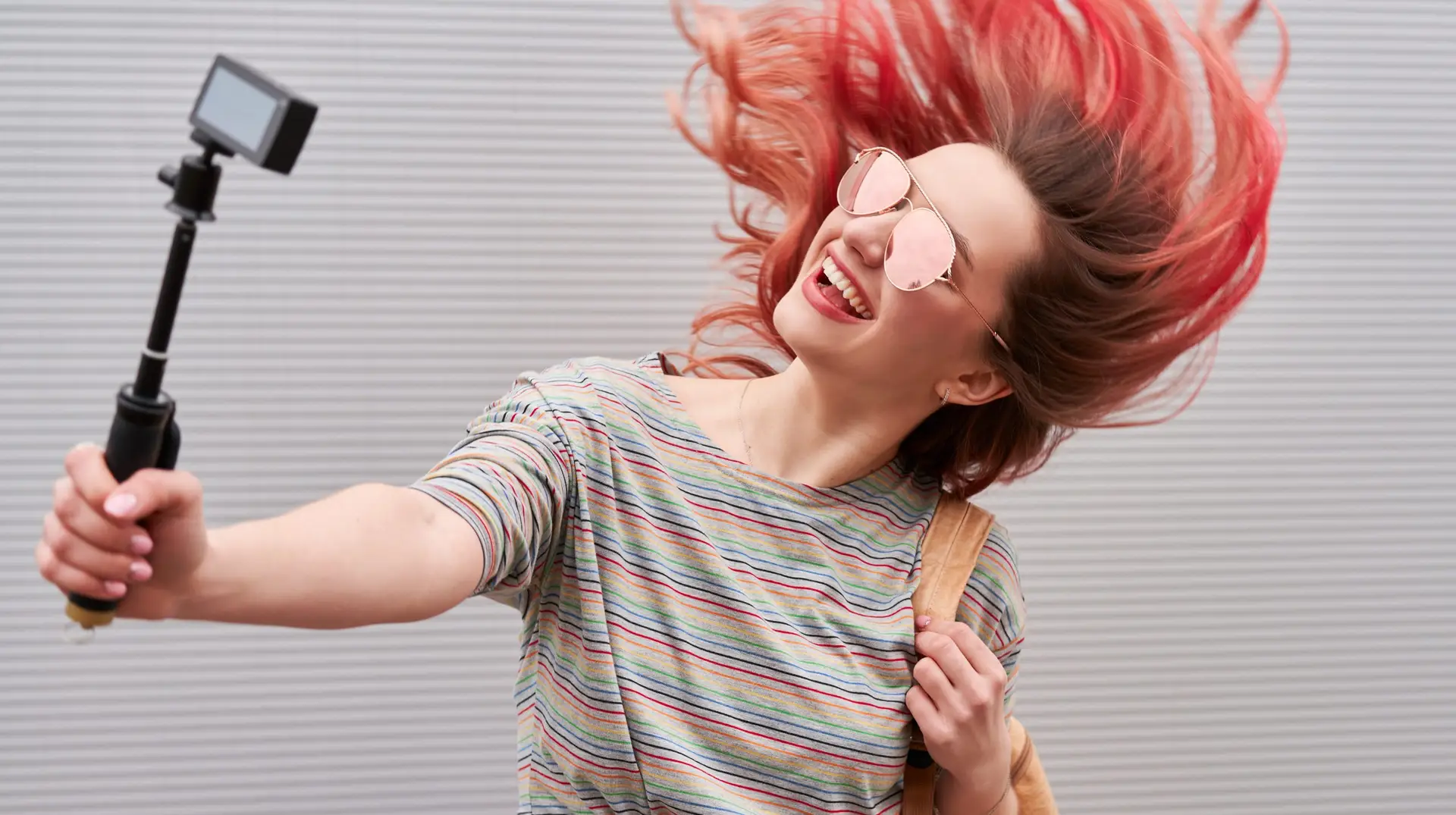 Young blogger woman with pink dyed flying hairs dancing near the grey wall and making vlog by action camera
