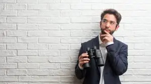 Young business man wearing a suit against a white bricks wall thinking and looking up, confused about an idea, would be trying to find a solution