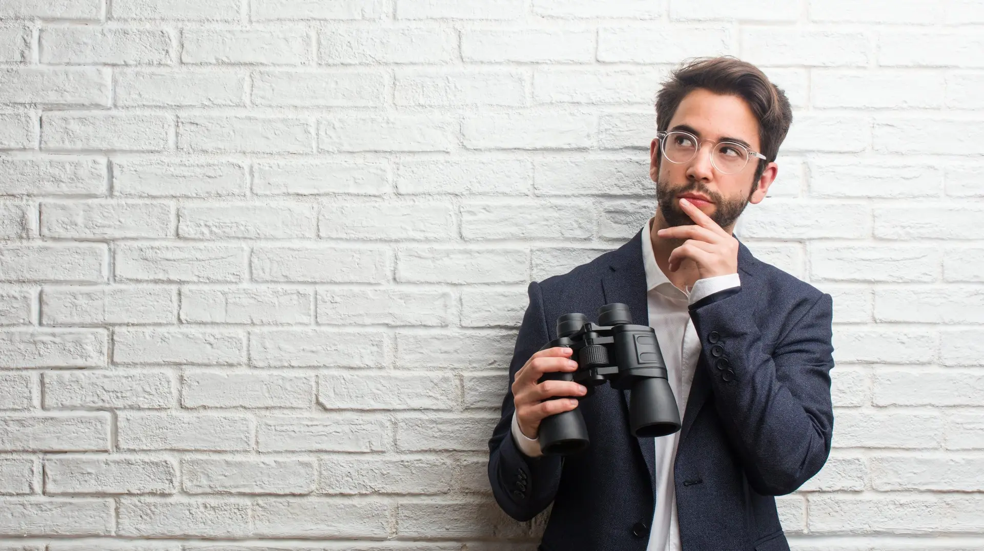 Young business man wearing a suit against a white bricks wall thinking and looking up, confused about an idea, would be trying to find a solution