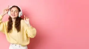 Beautiful stylish female model singing favorite song, listening to music in wireless headphones, smiling and dancing, standing over pink background
