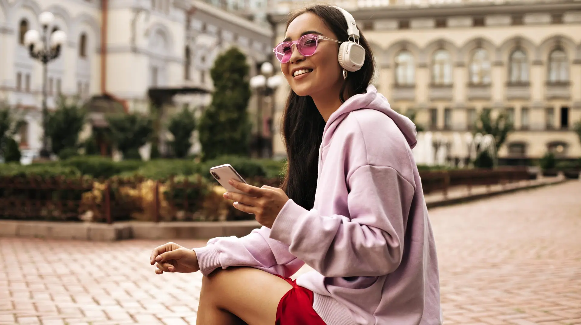 Charming brunette woman in pink hoodie and sunglasses looks away, holds phone and listens to music