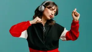 confident young caucasian woman wearing headphones keeping fists in air dancing with closed eyes isolated on blue background