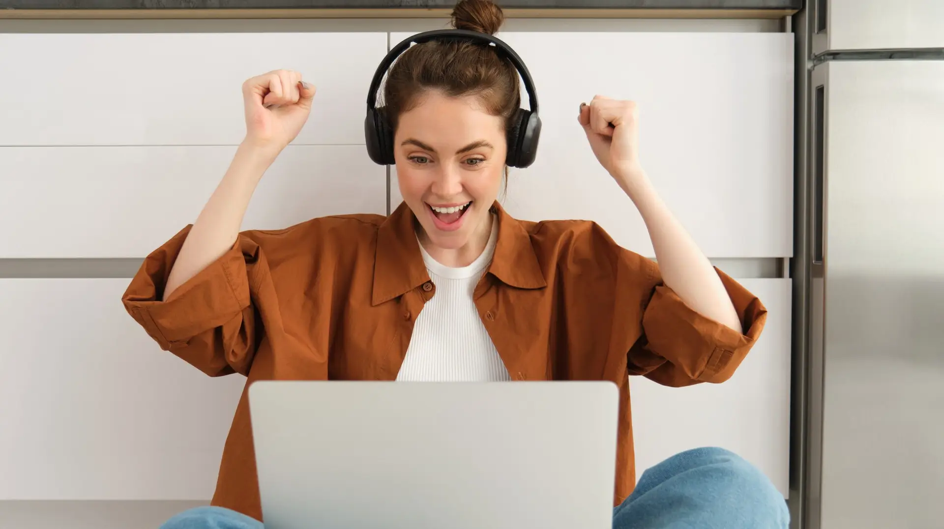 Excited young woman, programmer achieves goal, triumphing, winning on laptop, sitting in headphones, dancing with hands, celebrating victory