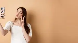 Fashion female blogger pucker lips and show v-sign at smartphone camera, taking selfie for social media, standing on beige background