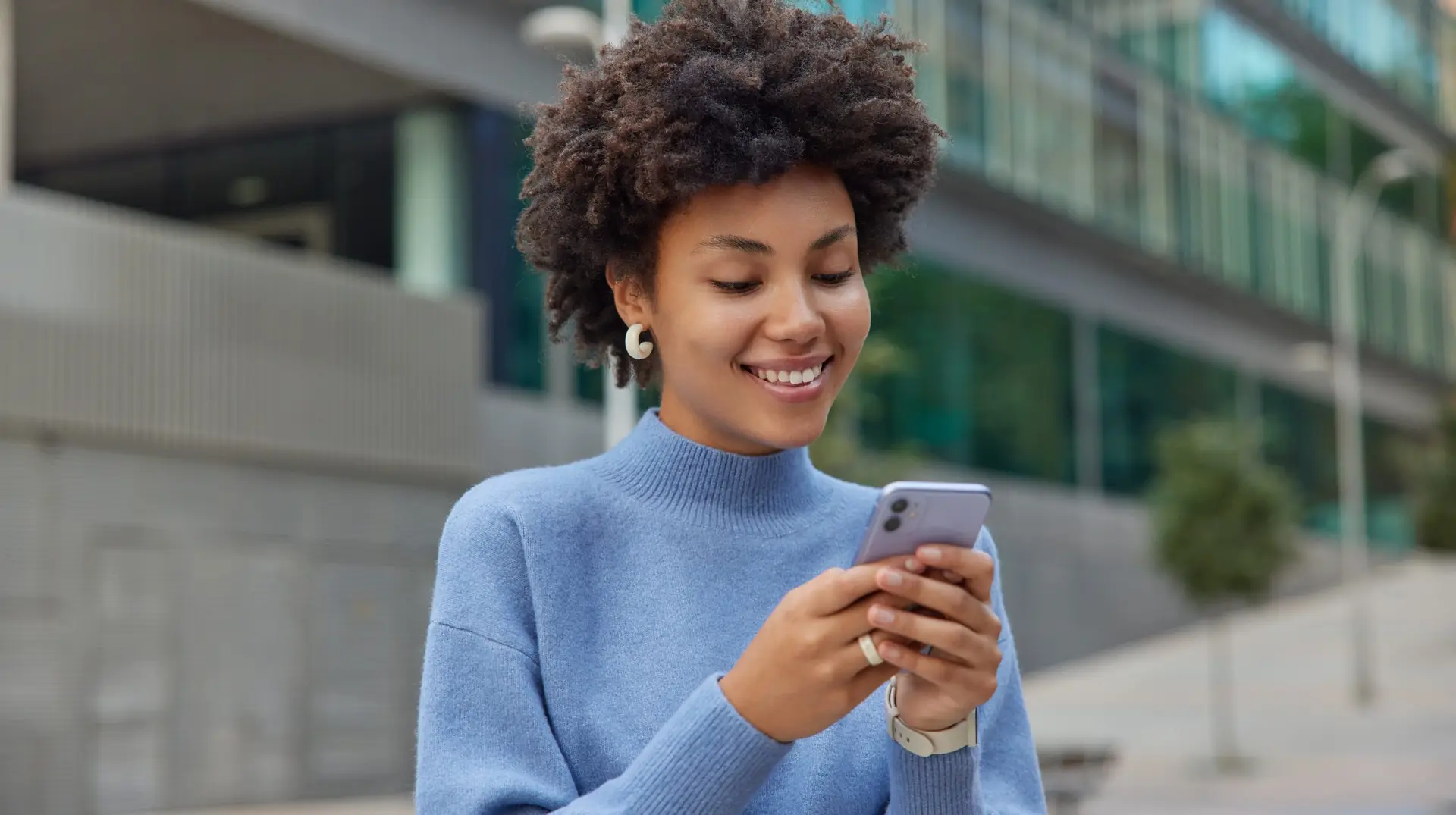 Glad young woman with curly bushy hair holds modern mobile phone downloads amazing application surfs social networks wears casual blue jumper smiles positively poses against blurred background