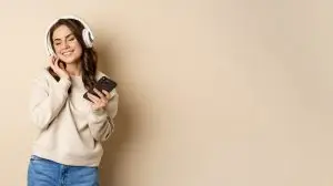 Happy caucasian woman listening music in headphones on smartphone app, dancing and having fun, standing over beige background