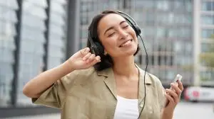 Happy girl dances on street and listens music in wireless headphones, holds mobile phone, using streaming app