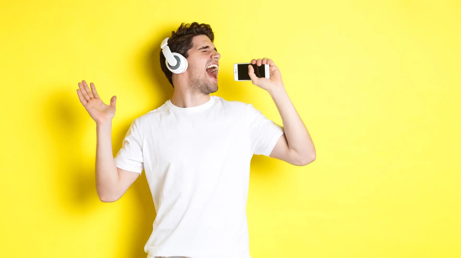 Happy guy playing karaoke app in headphones, singing into smartphone microphone, standing over yellow background
