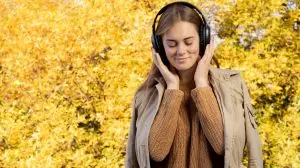 Happy woman standing on yellow autumn leaves background listening to music in headphones. Happy young girl listening to a podcast. Banner. Lifestyle, autumn fashion, beauty