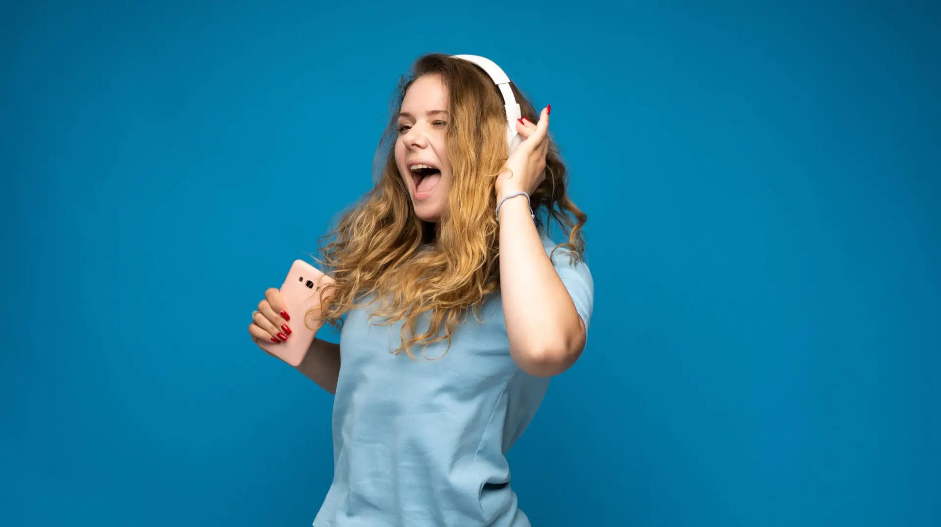 Happy young woman enjoying music in headphones with smartphone in hand on blue backdrop
