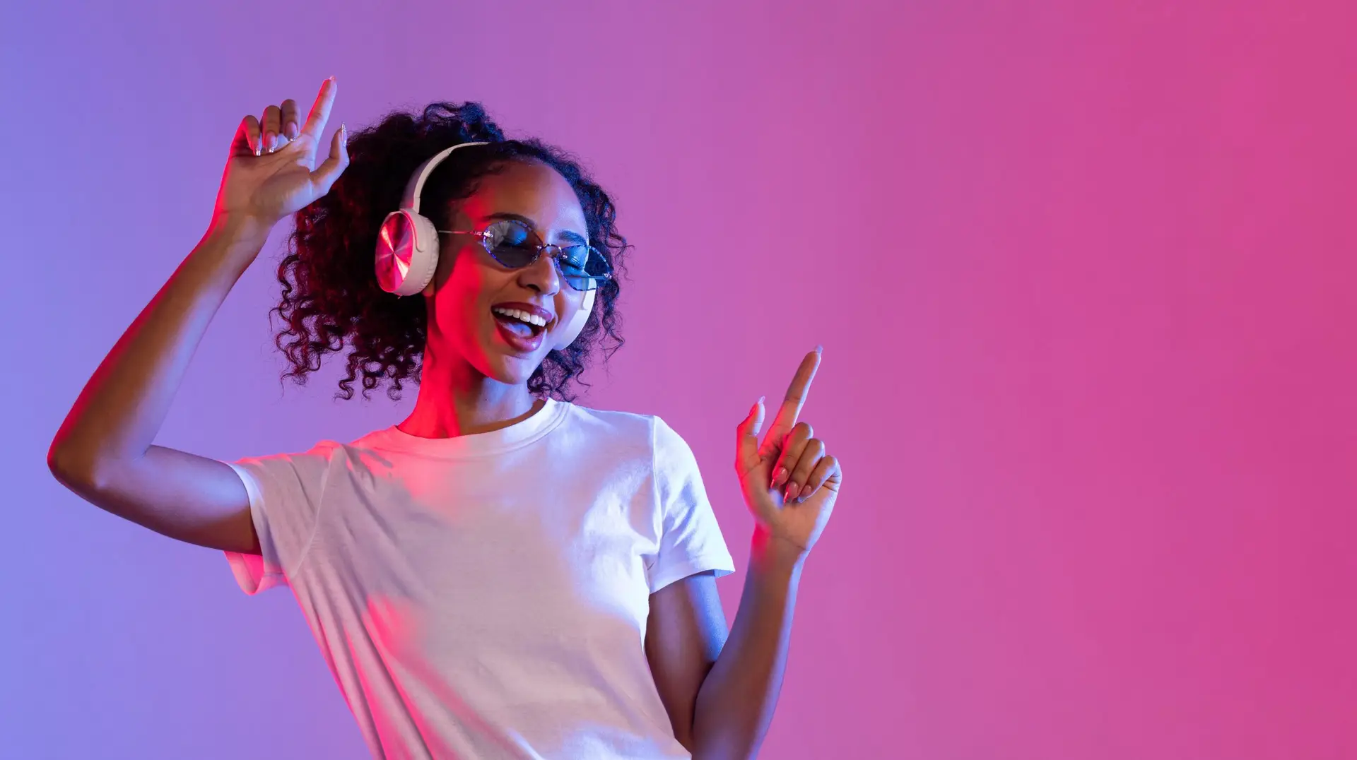 Joyful black woman with sunglasses and headphones dancing on neon background, free space