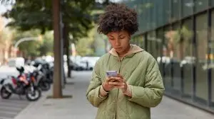 Photo of curly haired young woman in jacket uses mobile phone for chatting online strolls outdoors against blurred urban background holds modern device downloads application for finding route
