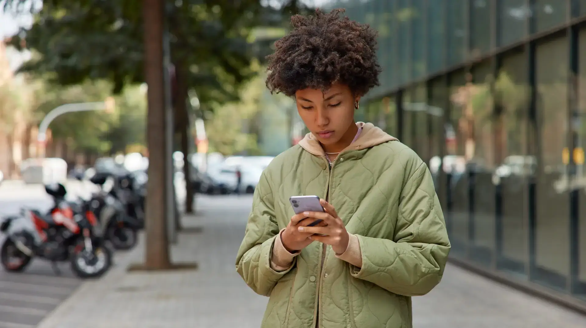 Photo of curly haired young woman in jacket uses mobile phone for chatting online strolls outdoors against blurred urban background holds modern device downloads application for finding route