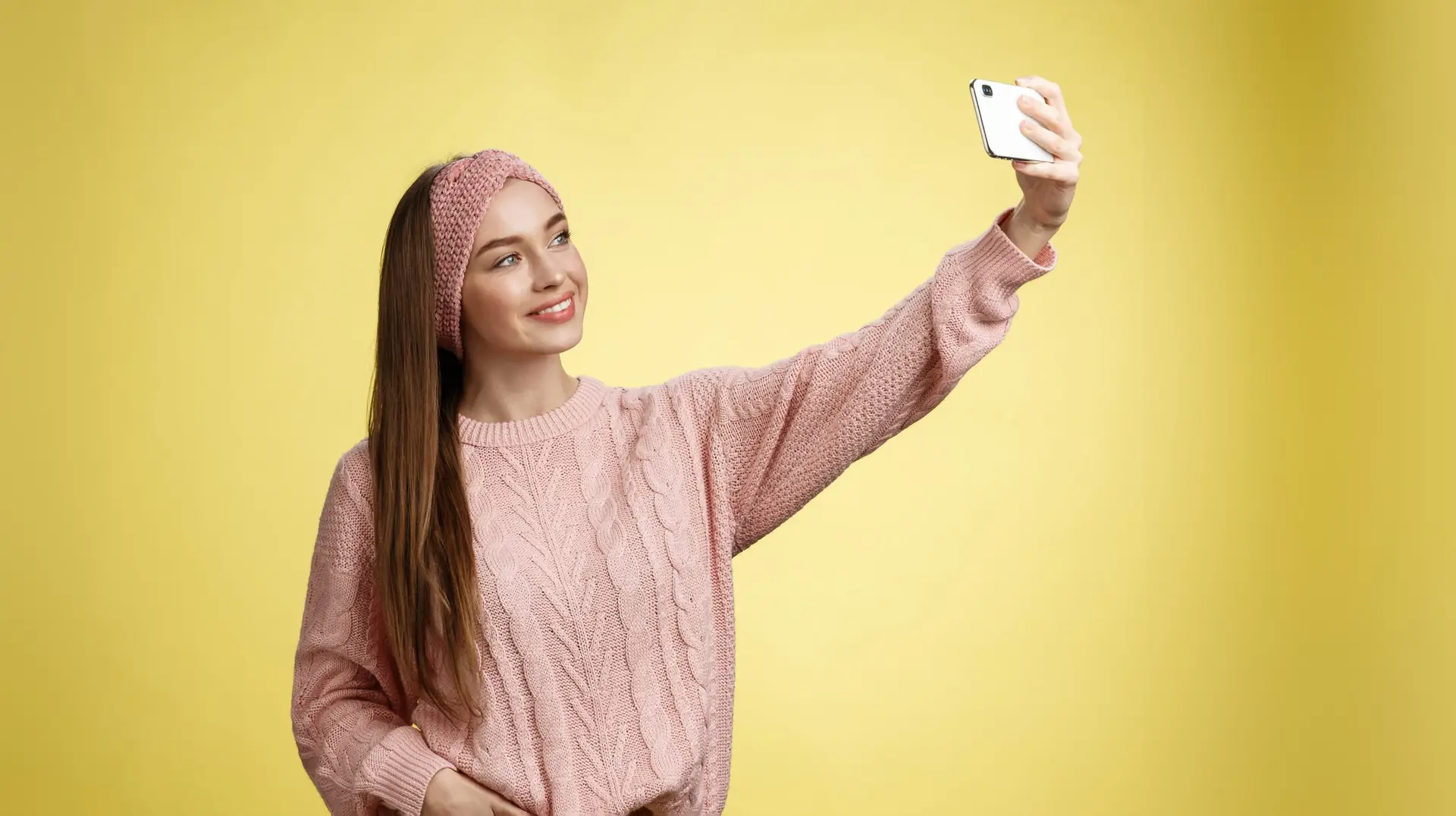 Popular glamour young female internet lifestyle blogger taking selfie on new smartphone extending arm taking picture herself against yellow background smiling at cellphone screen, posing cheeky