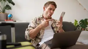 Portrait of a guy in living room, pointing at mobile phone, sitting with laptop and suitcase