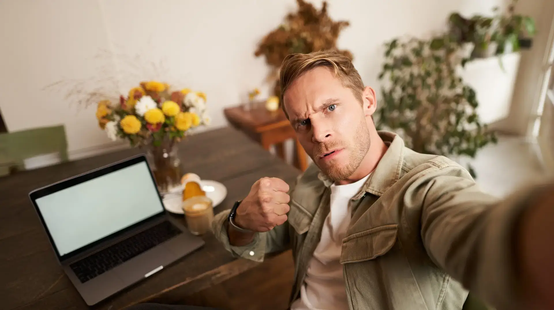 Portrait of serious man takes selfie with fist and threatening face, sits in cafe, poses in coffee shop in front of table with laptop and glass of coffee