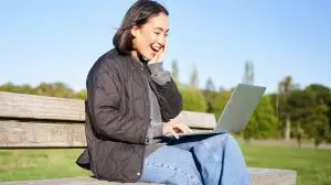 Portrait of smiling asian girl sits on bench in park, talks to friend online via laptop, video chats, using computer to study on remote