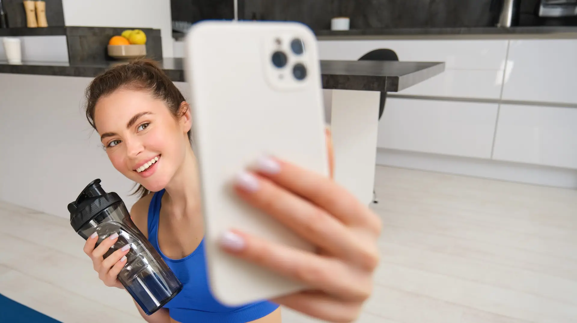 Portrait of sportswoman takes selfie with water bottle in living room, holds smartphone and poses for photo while doing fitness workout