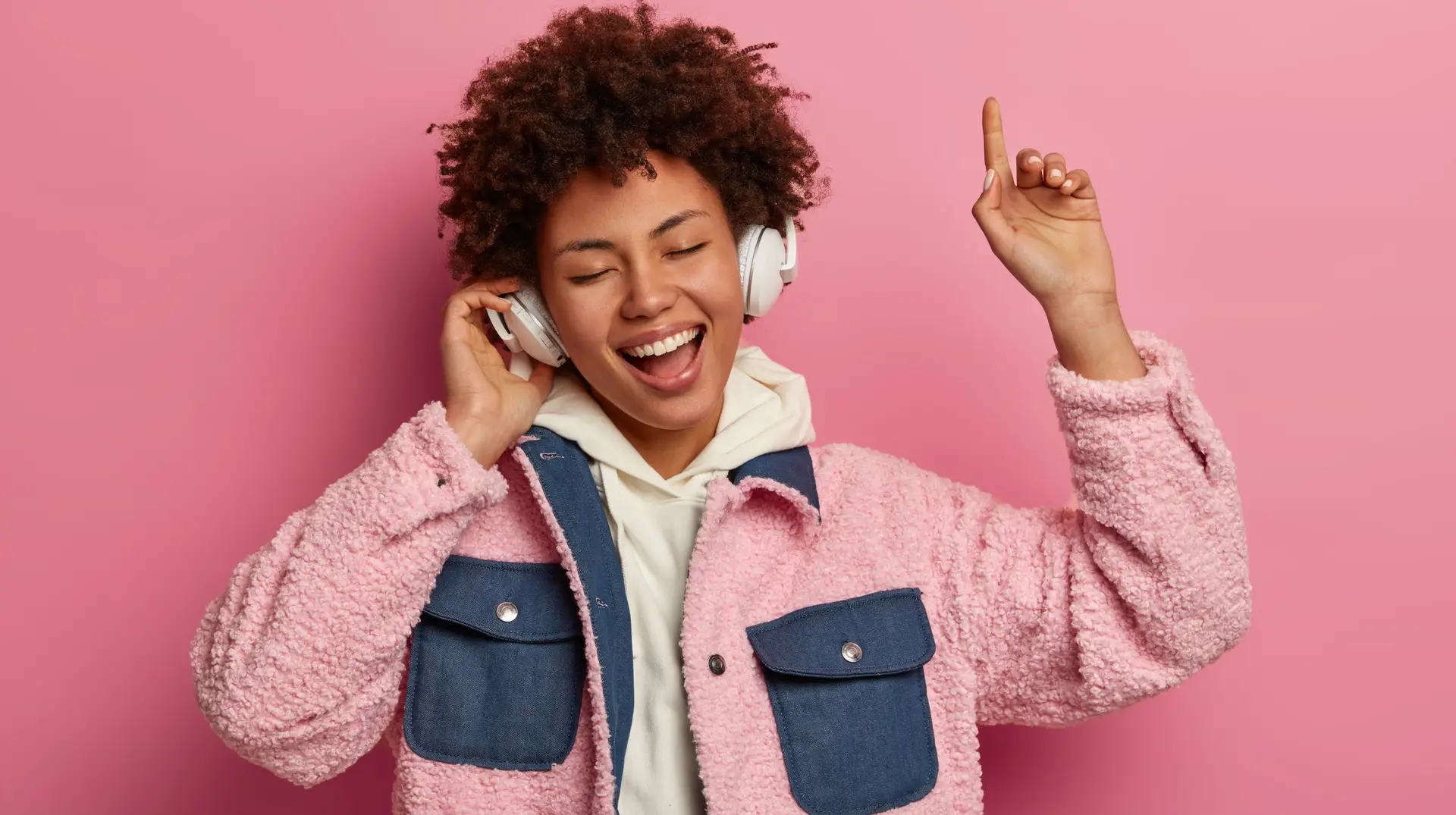 Happy relaxed ethnic female closes eyes and raises arm, dances joyfully to popular music, enjoys loud sound in headphones, listens new album of favorite band, moves to rhythm, isolated on pink