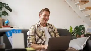 Portrait of young handsome man sitting with laptop, has passport and two plane tickets prepared, planning his journey on computer, booking hotels and restaurants online for the trip