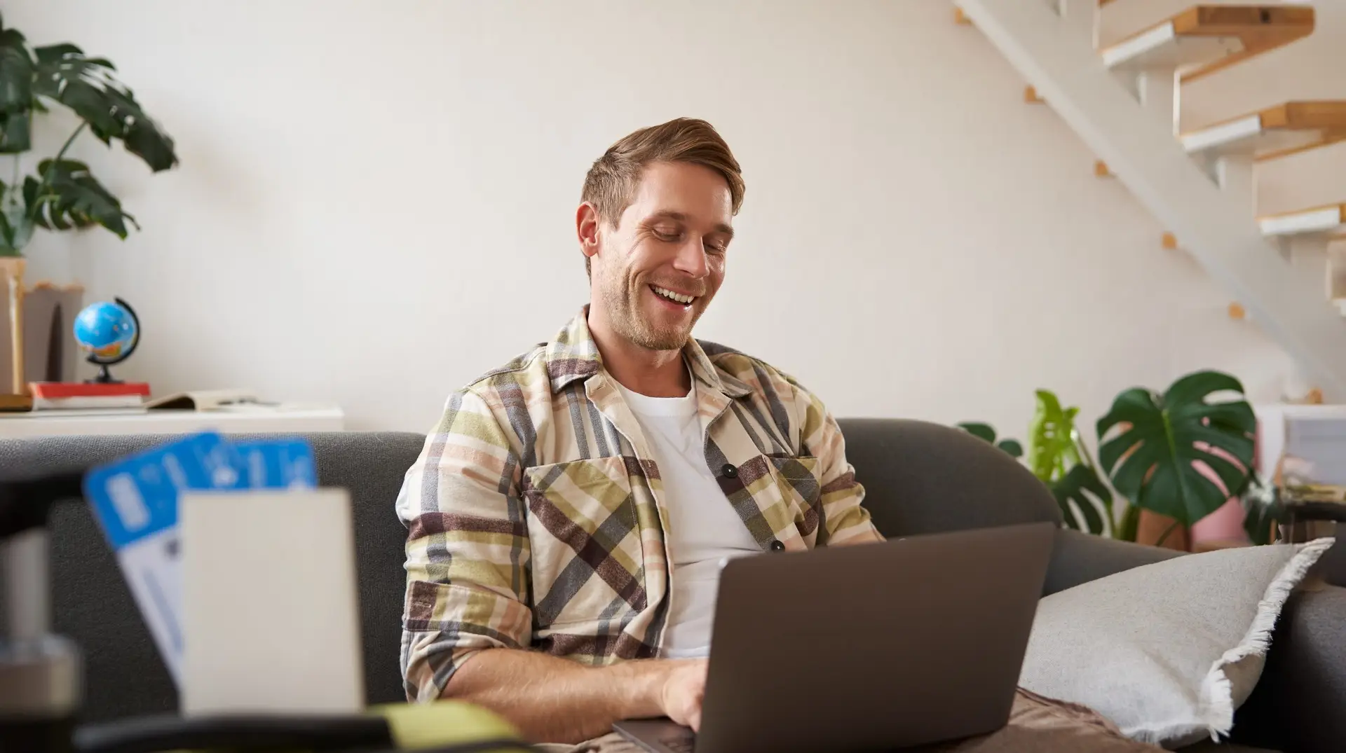 Portrait of young handsome man sitting with laptop, has passport and two plane tickets prepared, planning his journey on computer, booking hotels and restaurants online for the trip