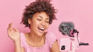 Positive pretty brunette Afro American female blogger focused at smartphone on tripod makes online streaming has own channel smiles gladfully poses against pink background. Vlogging concept.
