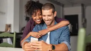 Smiling,Young,Couple,Embracing,While,Looking,At,Smartphone.,Multiethnic,Couple