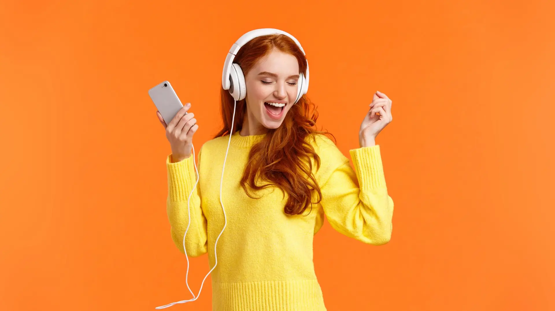 Technology, emotions and gadgets concept. Cheerful good-looking redhead woman dancing with hands up, closed eyes, smiling joyfully, enjoy awesome music sound in headphones, hold smartphone