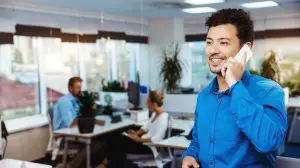 Young successful businessman speaking on phone, smiling, over office background.