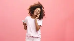 Getting rid of worried with good tunes in ears. Pleased attractive young african woman in trendy outfit listening music in wireless earphones touching earbud smiling dancing and holding smartphone