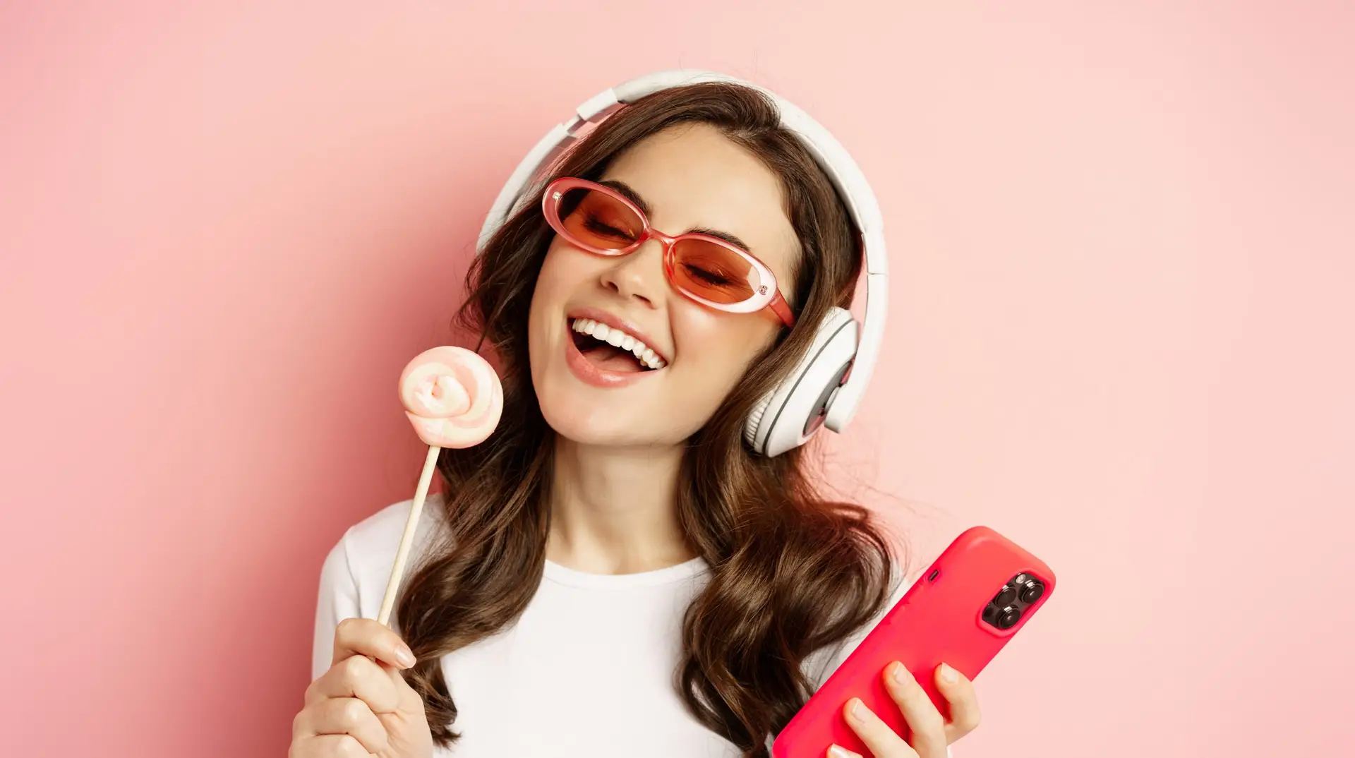 Beautiful female model listening music in headphones, holding lolipop and mobile phone, posing in sunglasses, standing over pink background