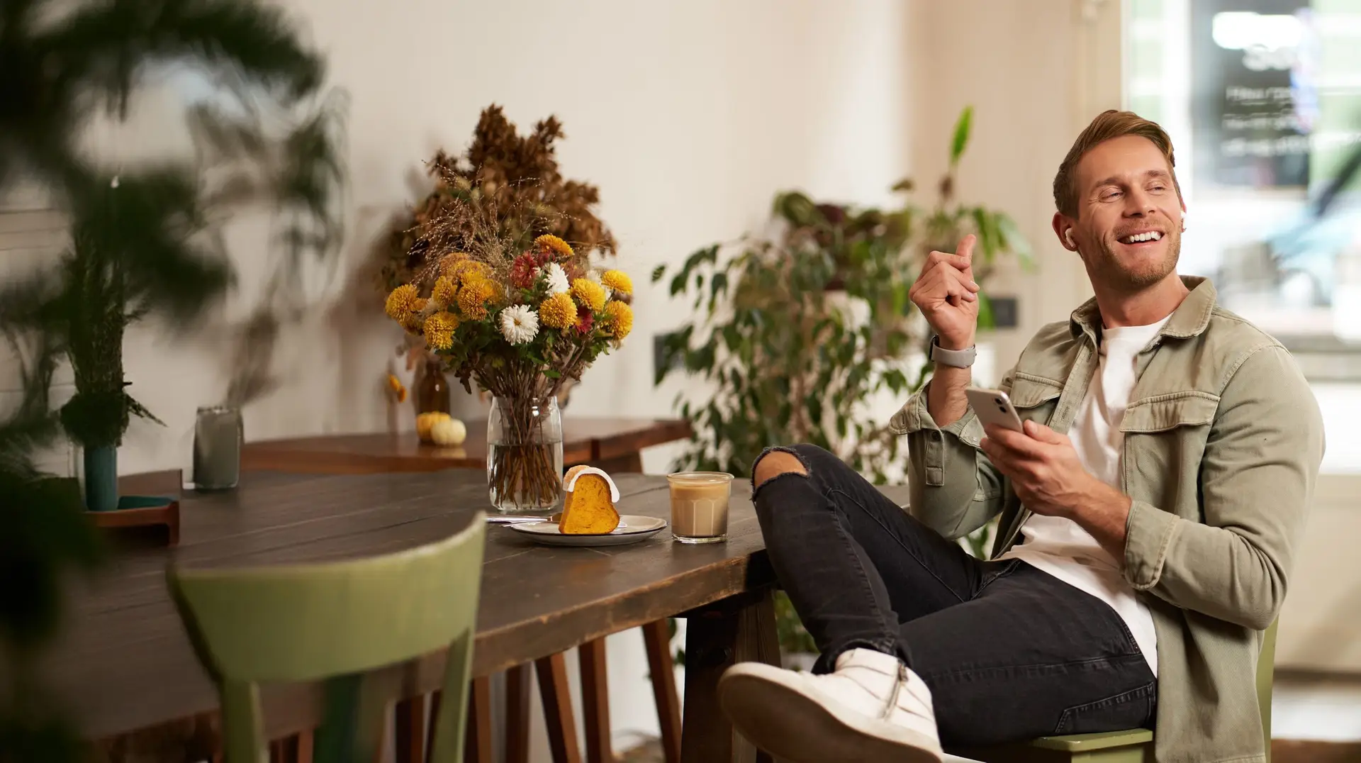 Portrait of young handsome man in casual clothes, sits in cafe and enjoys favourite song, listens to music in wireless headphones, vibing to the relaxing tunes in earphones, holding smartphone