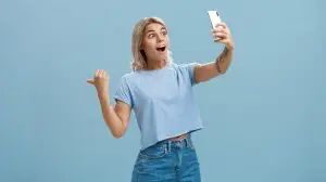 Girl recording video showing followers awesome scene attending cool event holding smartphone looking impressed and surprised at device screen pointing backwards indicating right over blue wall