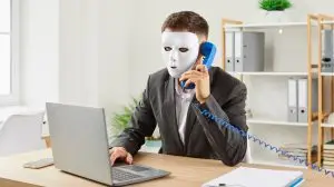 Anonymous,Man,Disguised,In,White,Face,Mask,Sitting,At,Office