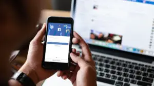 A person is holding a phone with the Facebook login page up, in the background, there is a laptop with the Facebook page open.