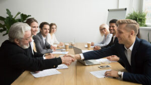A group of business partners are happily watching the two shaking hands, finalizing the negotiations.
