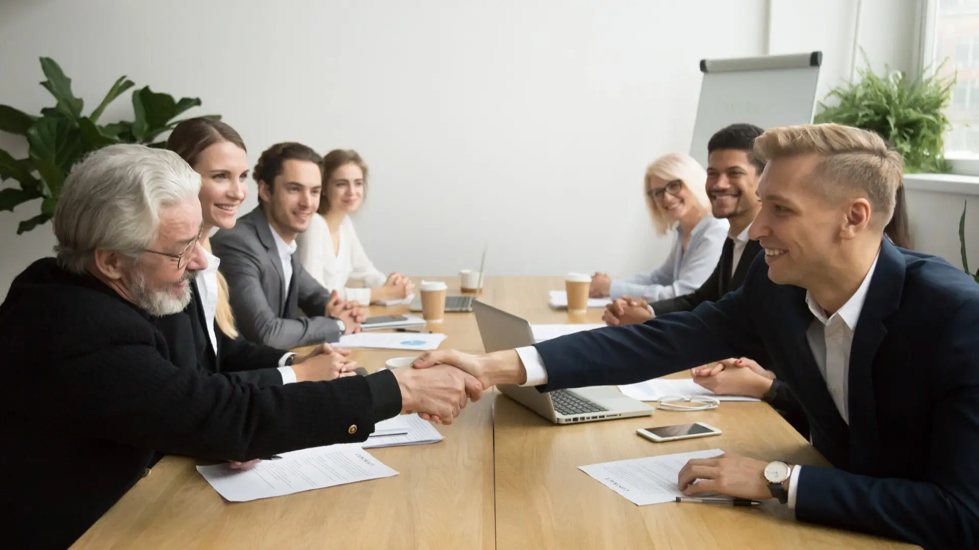 A group of business partners are happily watching the two shaking hands, finalizing the negotiations.