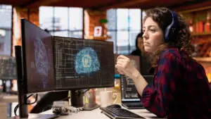 A woman is sitting in front of a double-screen computer, coding.