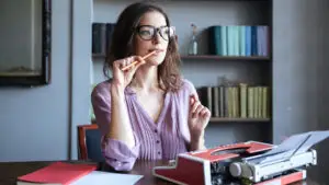 A woman sits behind a desk with a typewriter and a pencil, thinking.