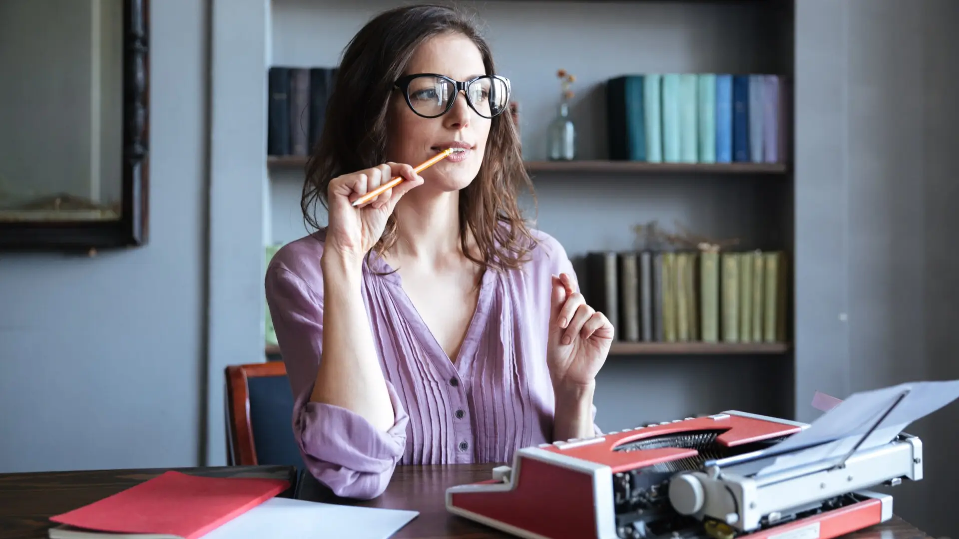 A woman sits behind a desk with a typewriter and a pencil, thinking.