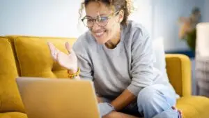 Modern happy young woman smile and have fun in video call computer online activity. Doing a live coaching call using internet and laptop connection. Smart female people sitting on yellow sofa with notebook pc
