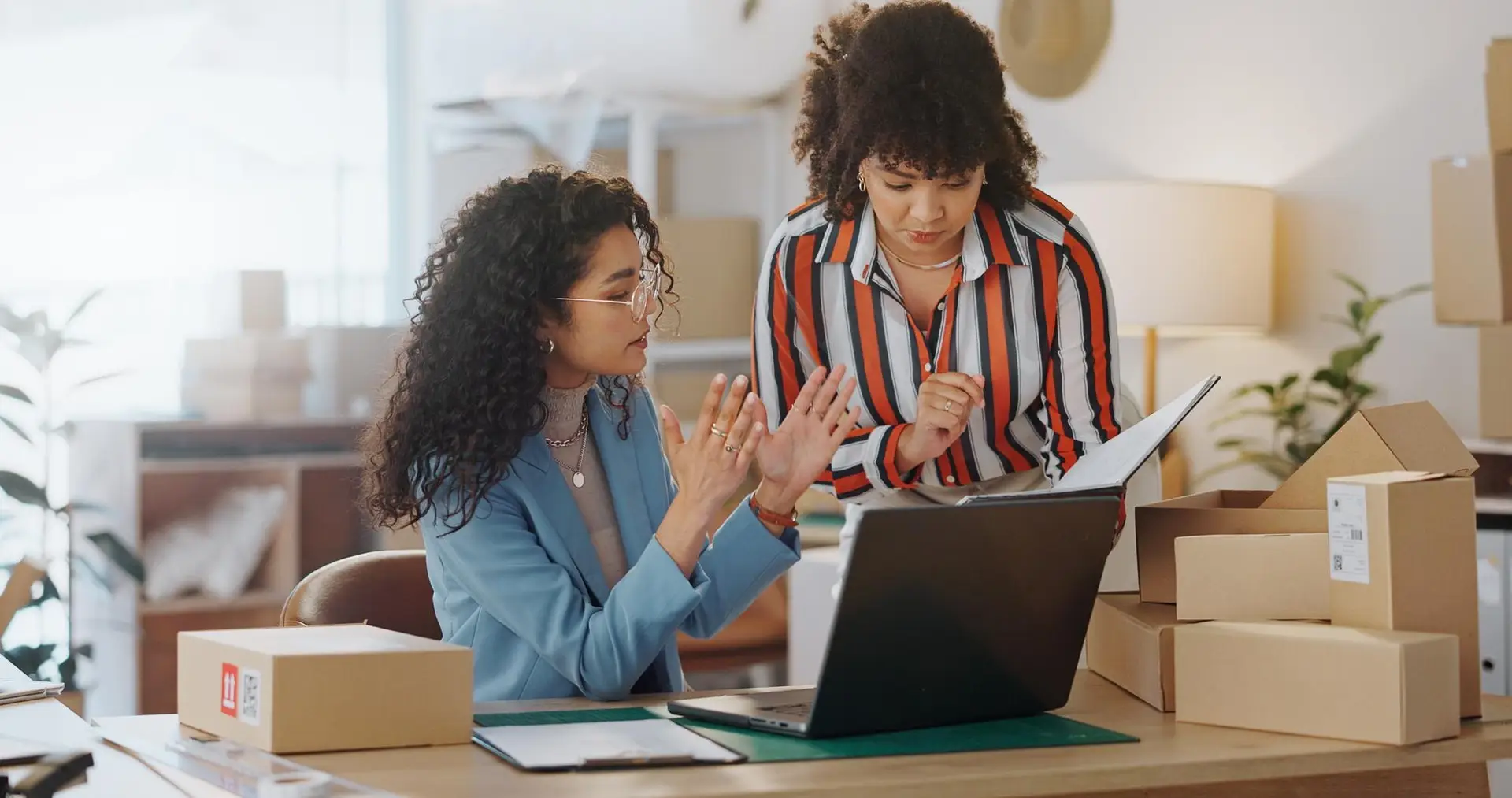 Two women are behind a computer surrounded with boxes while discussing how to build a business.