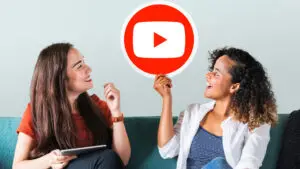 Two women are sitting on a couch, one of whom is holding a YouTube logo sign, and they're both looking at it.