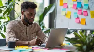 Digital marketer man sitting behind a table working on his computer, with sticky notes visible on a board next to him