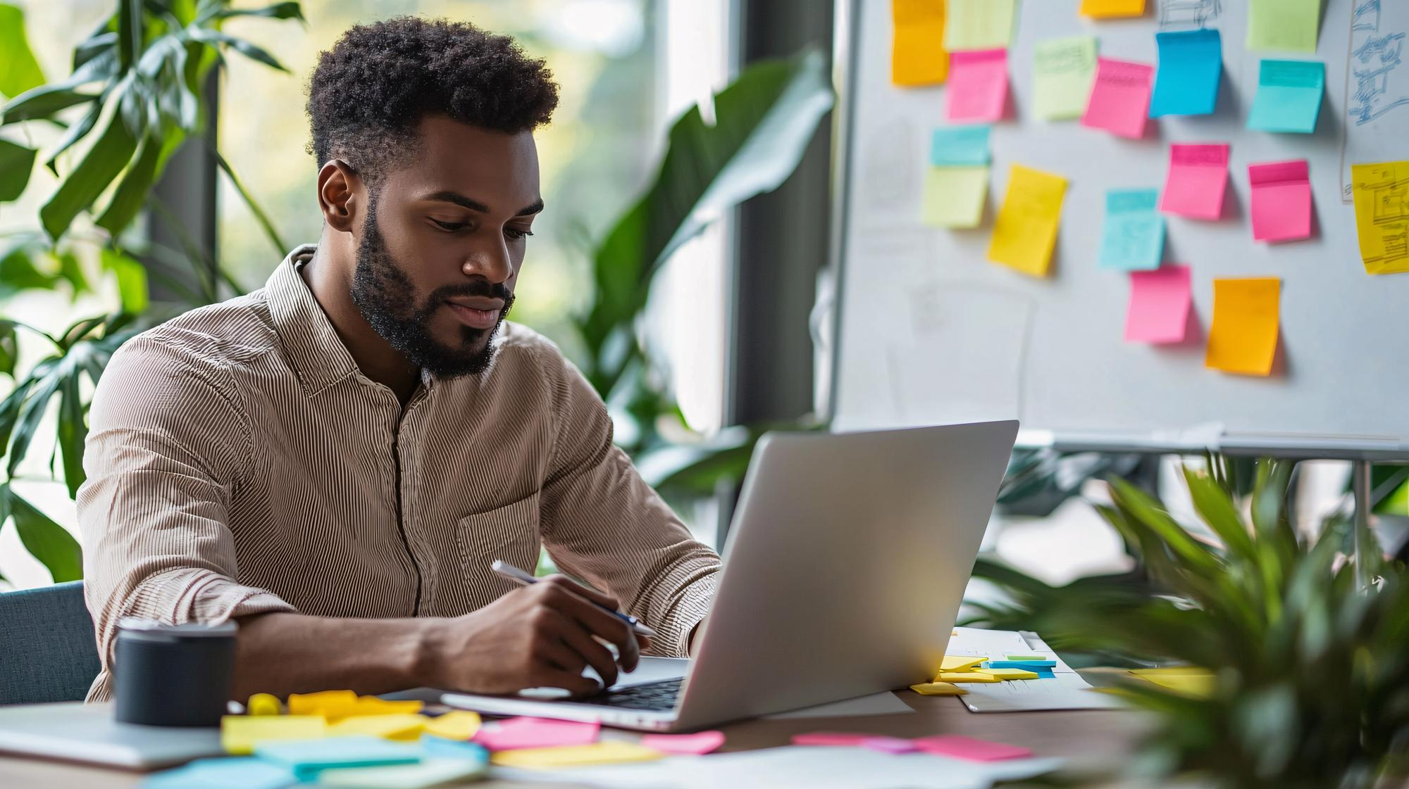 Digital marketer man sitting behind a table working on his computer, with sticky notes visible on a board next to him